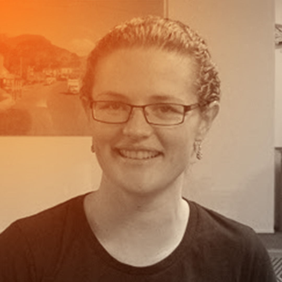 A pakeha woman with glasses, smiling in front of a red Summer of Tech banner