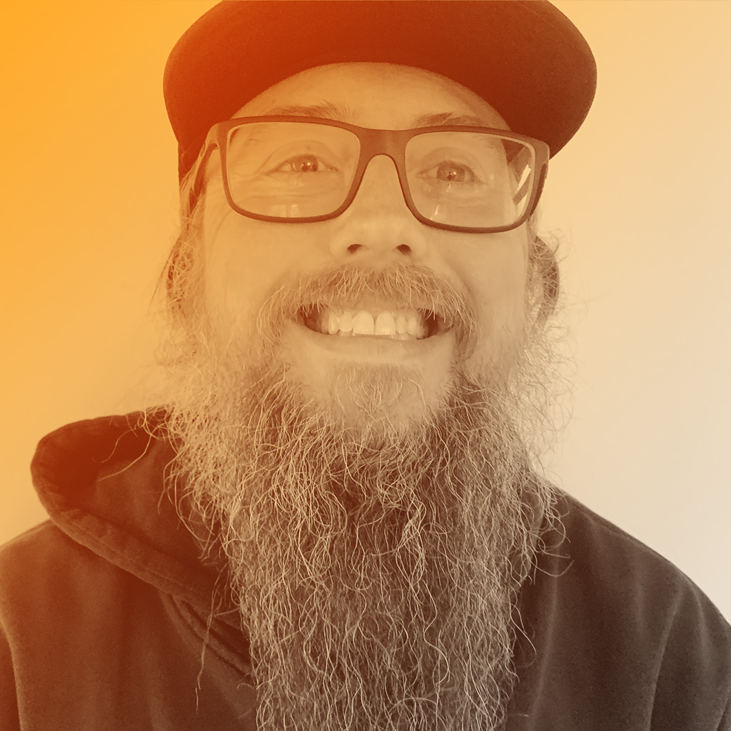 A middle aged white man with dark framed glasses, a baseball cap and a long beard, smiling.