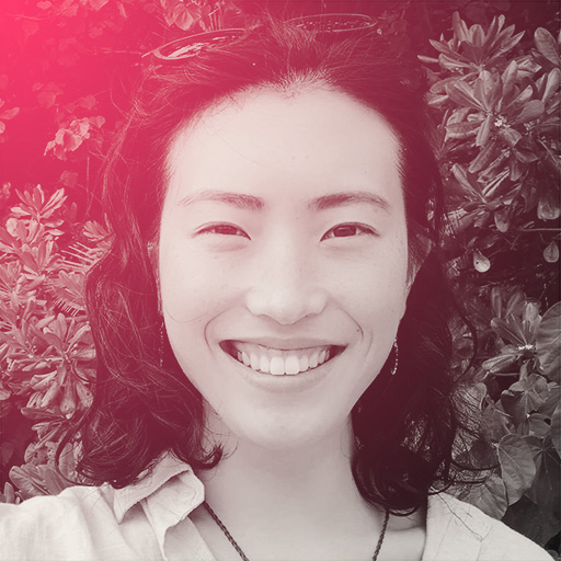 A portrait photo of Jenny smiling at the camera. She is wearing sunnies on her head, silver dangly earrings, a collared shirt, and is standing in front of some jungly bush.