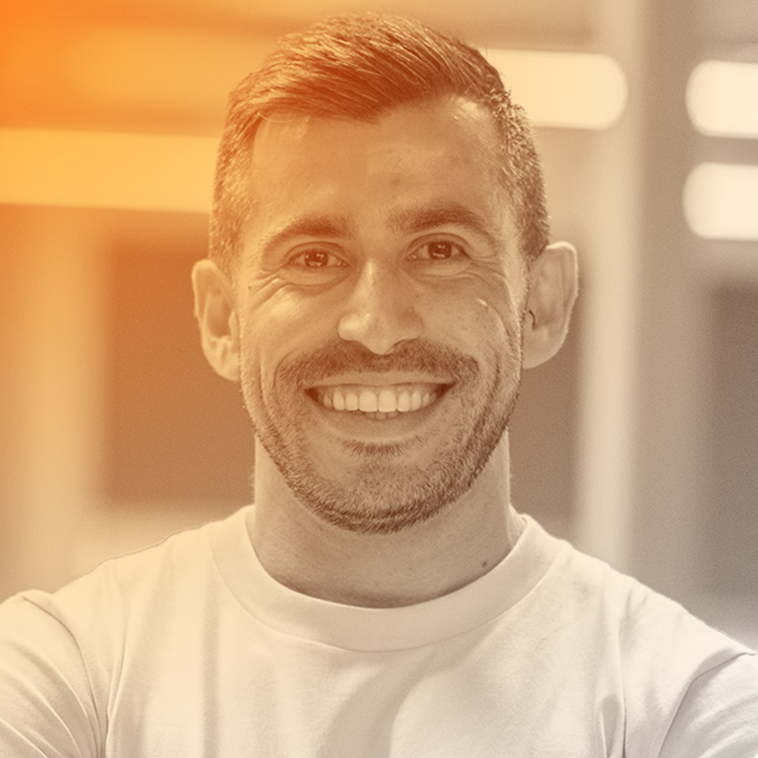 Smiling man with short dark hair and stubble, wearing a pale pink t-shirt, standing with arms crossed in an office setting.